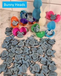 sixty-four blue stained wooden laser-engraved bunny head-shaped Easter egg reward tokens spread across a marble surface beside colorful open plastic Easter eggs, with rewards including build a blanket fort, family movie night, dance party, go bowling, and choice of reward