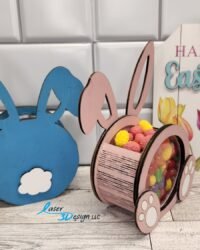 Blue and pink wooden bunny candy holders displayed side by side on a wood surface, with the pink bunny filled with colorful yellow and red candy visible through the clear acrylic belly, beside a Happy Easter decorative sign with tulips